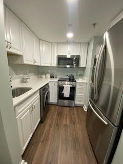 A modern kitchen with white cabinets, stainless steel appliances, and a dark wooden floor. It features a refrigerator, oven, microwave, and a sink. The counter tops are white, and there is a small area with various kitchen items.