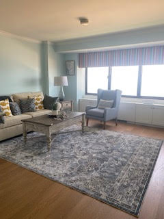 A cozy living room with light blue walls features a beige sofa with patterned cushions, a gray armchair, and a wooden coffee table atop a patterned rug. A striped valance adorns the window, and a lamp stands beside the sofa. Natural light fills the space.