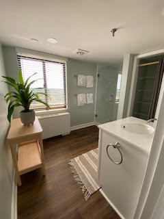 A modern bathroom with a light wood floor, a white vanity with a towel ring, and a potted plant on a wooden stand. A window with blinds allows natural light in, and white towels hang near a frosted glass shower door. A striped rug lies on the floor.