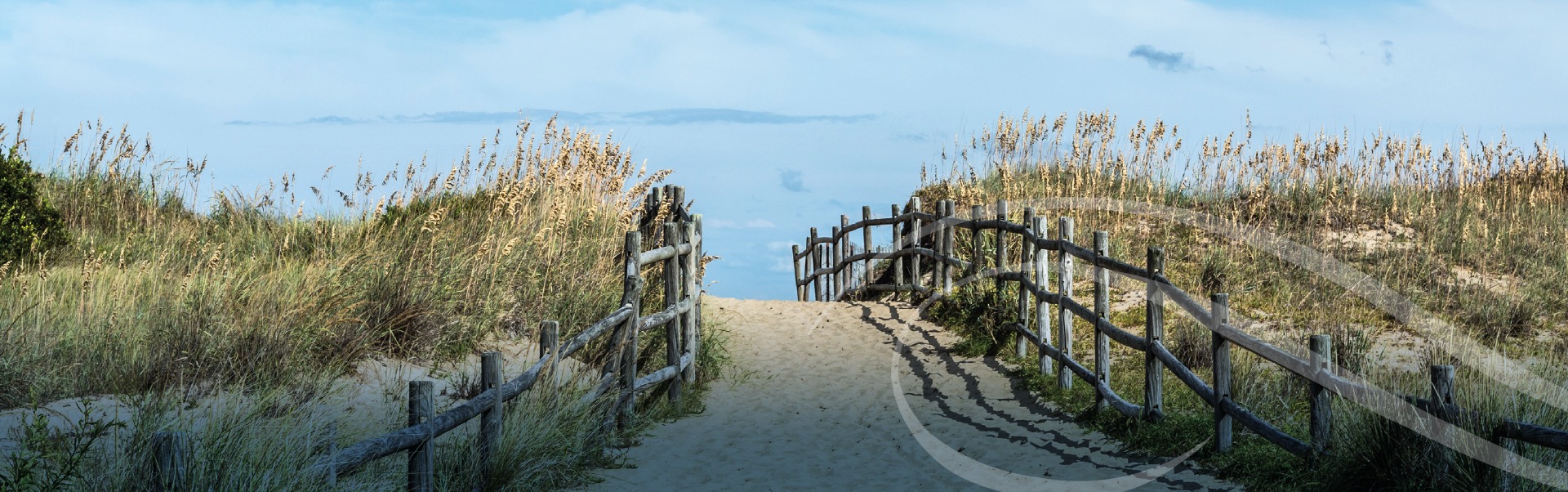 A sandy path leads through tall grass and wooden fences toward a beach with a clear blue sky in the background. Waves gently roll up to the distant shoreline, enhancing the serene and inviting atmosphere.