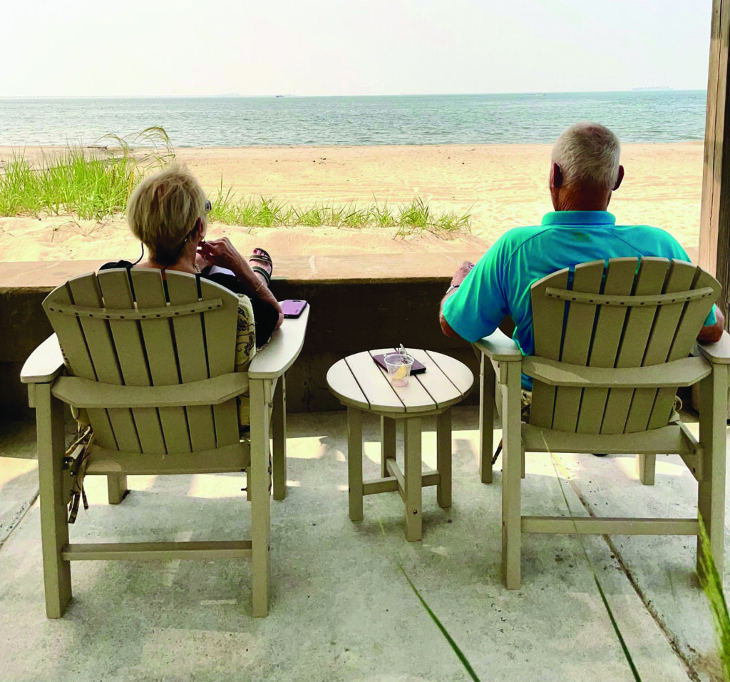 Two people sit in Adirondack chairs on a patio facing a tranquil beach. The sand and ocean stretch out under a hazy sky. A small table between them holds a drink, while grass peeks in from the side.