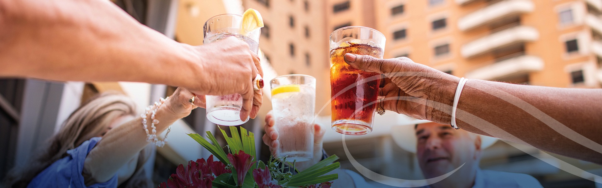 People raising glasses for a toast with drinks that include lemon slices and a cocktail. The scene is outdoors, with a city building in the background and a vase of red flowers nearby.