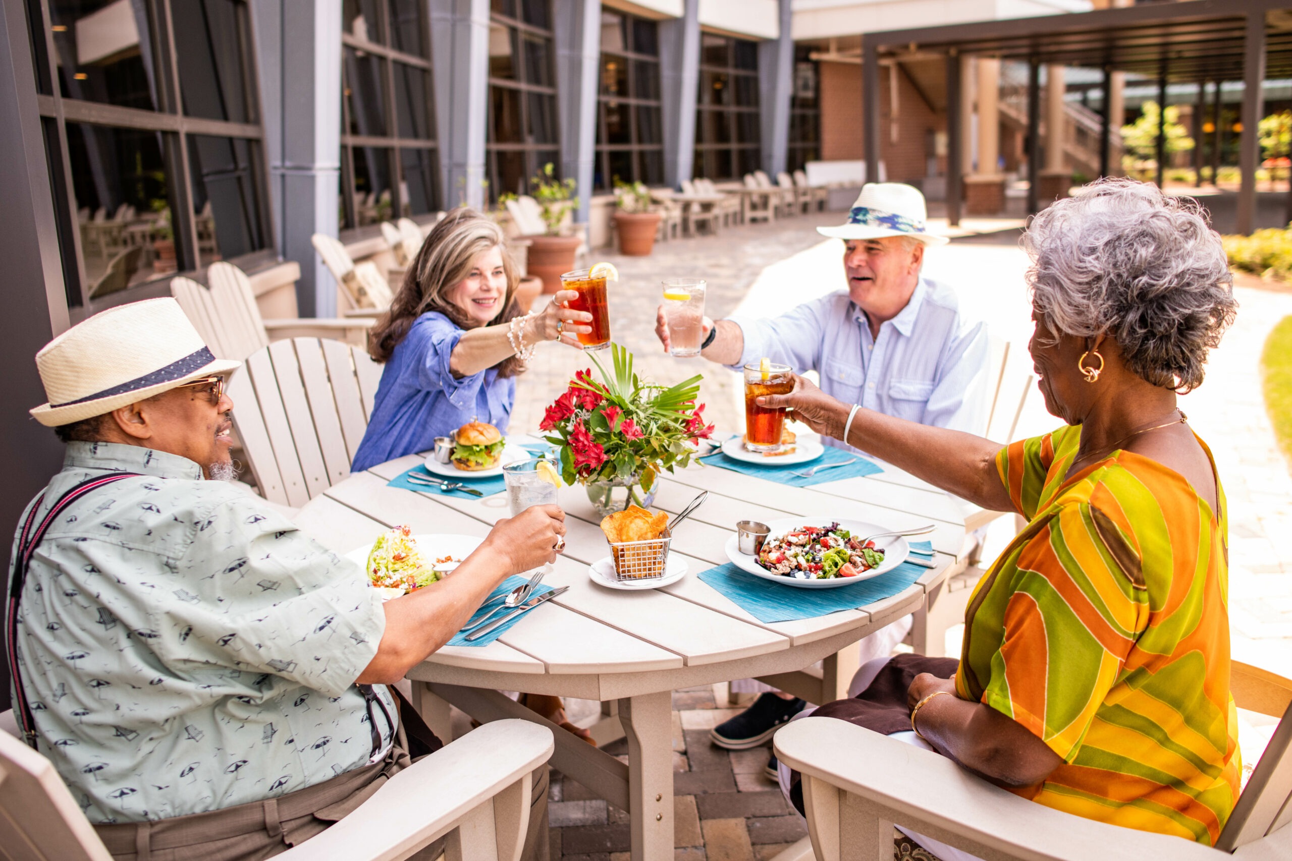 Four people sitting around an outdoor table toasting with drinks. They have plates of food in front of them. The setting is a sunny patio with flowers on the table and a modern building in the background.