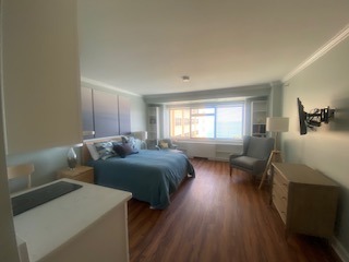 A cozy bedroom with a large bed covered in blue bedding, a soft armchair in the corner, a dresser with a lamp, and a wall-mounted TV. The room has wooden flooring and a large window letting in natural light.