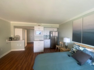 A cozy bedroom with green walls and wooden floors features a bed with blue bedding and decorative pillows. A small kitchen area with a fridge and microwave is visible in the background. A large ocean-themed painting hangs on the wall.