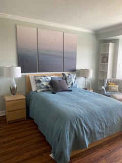 A cozy bedroom with a wooden bed, teal bedding, and geometric pillows. Two bedside tables with lamps flank the bed. A large triptych of a serene ocean scene hangs above. A patterned chair sits to the right, with a built-in shelf beside it.