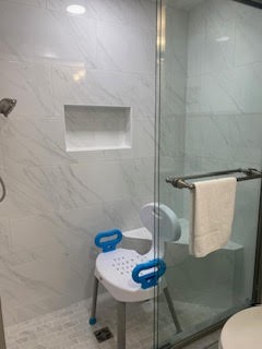 A bathroom shower with white tiled walls and a glass door. Inside, there is a white shower chair with blue handles and a backrest, positioned next to a wall-mounted grab bar with a white towel hanging over it.