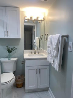 A modern bathroom with light gray walls features a white vanity with a sink and a large mirror above, illuminated by wall lights. A toilet with a wicker basket is nearby, and white towels hang on a bar next to the sink. Marble-tiled flooring adds elegance.