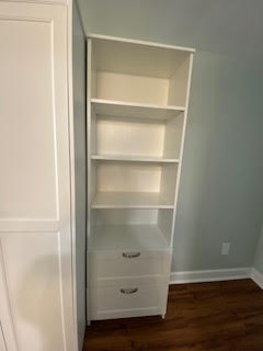 A tall white bookshelf with five shelves and two drawers at the bottom stands against a light gray wall. The floor is wooden, and the bookshelf is adjacent to a white cabinet.