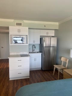 A small kitchen area featuring white cabinets, a stainless steel refrigerator, a microwave above the counter, and a sink. A wooden chair sits beside the counter. The flooring is wooden, and part of a bed with a blue cover is visible.