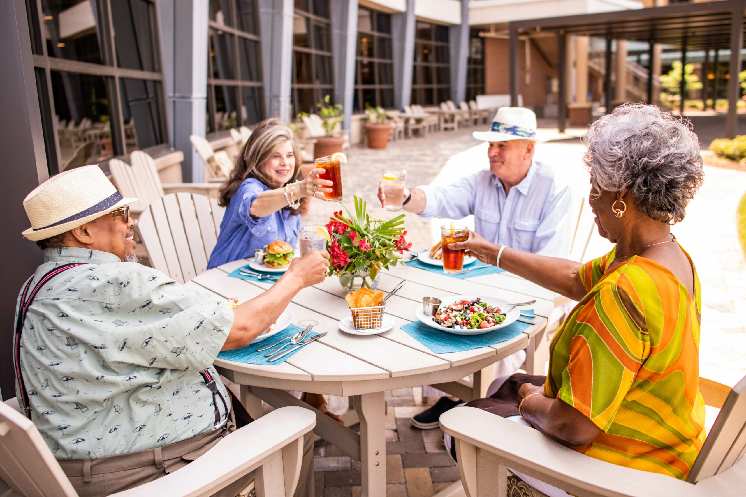 Four people sit around an outdoor table, smiling and raising glasses in a toast. The table is set with plates of food and a floral centerpiece. Its a sunny day with a building in the background.