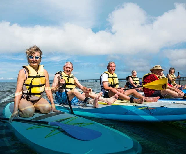 A group of elderly people sitting on paddleboards in the water, wearing life vests and smiling. The sky is partly cloudy, and a pier is visible in the background.