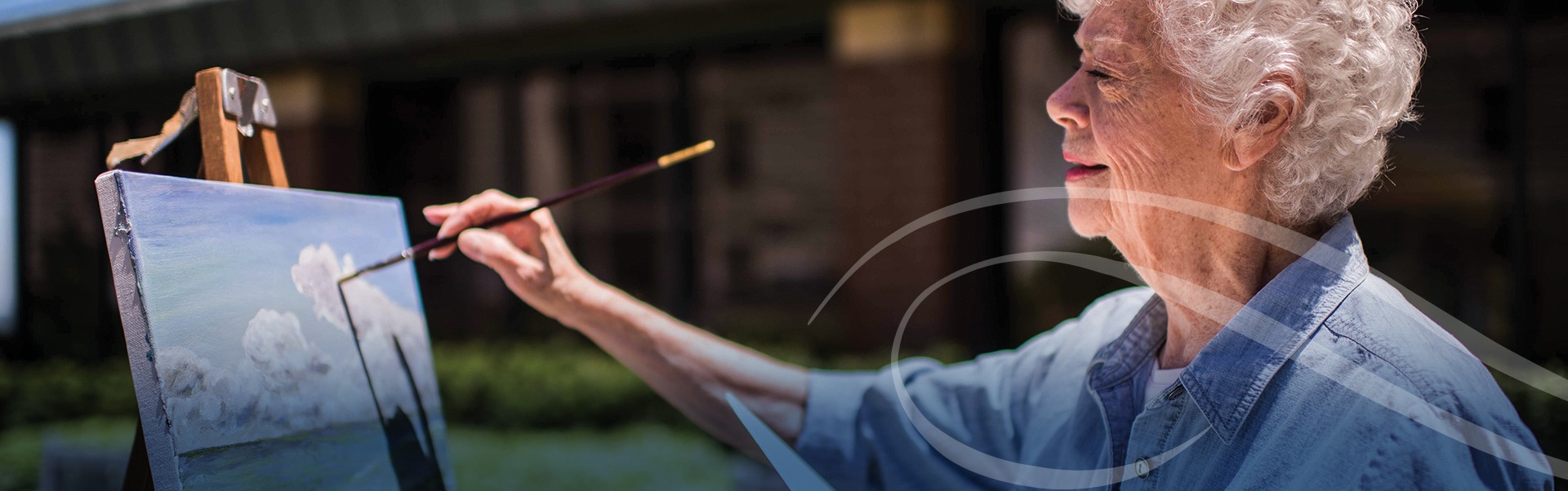 Elderly woman with curly white hair, wearing a blue shirt, painting a landscape on a canvas outdoors. She is focused, holding a brush against a partly cloudy sky scene.