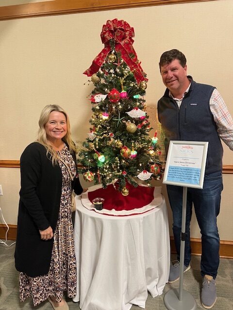 A woman and a man stand smiling beside a decorated Christmas tree on a table. The tree is adorned with colorful ornaments and a red bow on top. A framed sign is placed in front of the tree. The setting appears festive.