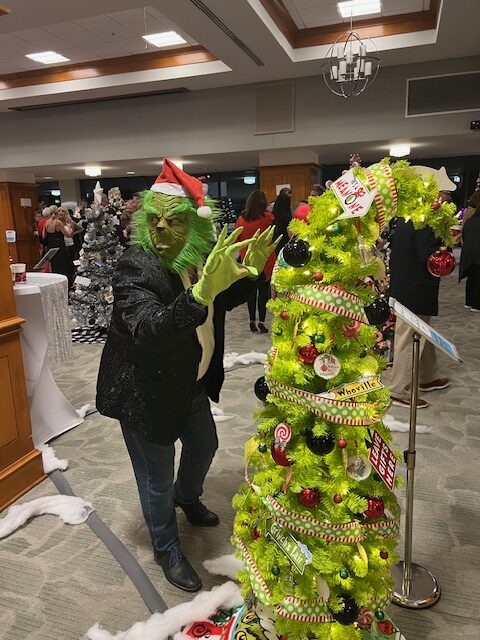 Person in a Grinch costume playfully posing with a whimsical green Christmas tree decorated with red and black ornaments, candy-themed accents, and a crooked top. The scene takes place indoors with other people and decorated trees in the background.