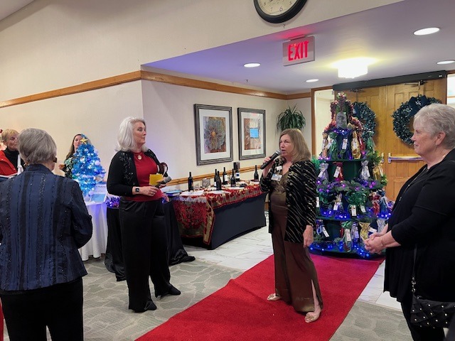 A group of elegantly dressed women gather in a decorated room with holiday trees and a red carpet. One woman speaks into a microphone. Tables with glasses and tablecloths are in the background, and a clock is on the wall above.