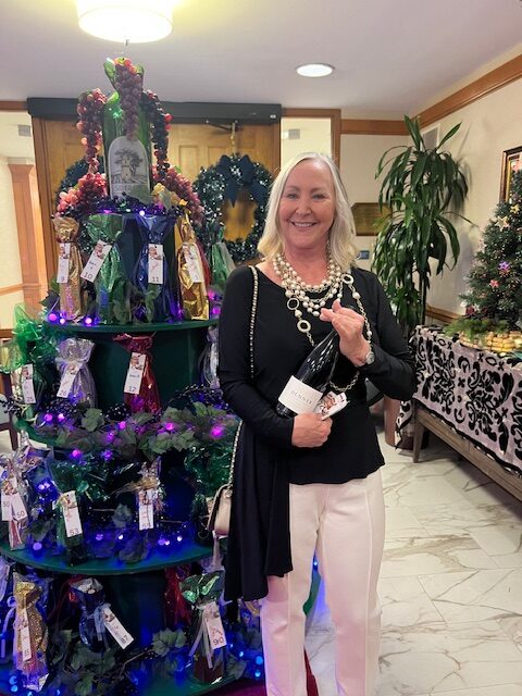 A smiling woman stands indoors holding a large bottle of wine. She is next to a decorative Christmas display with wrapped gifts and festive lighting. There are holiday decorations, including a wreath, in the background.