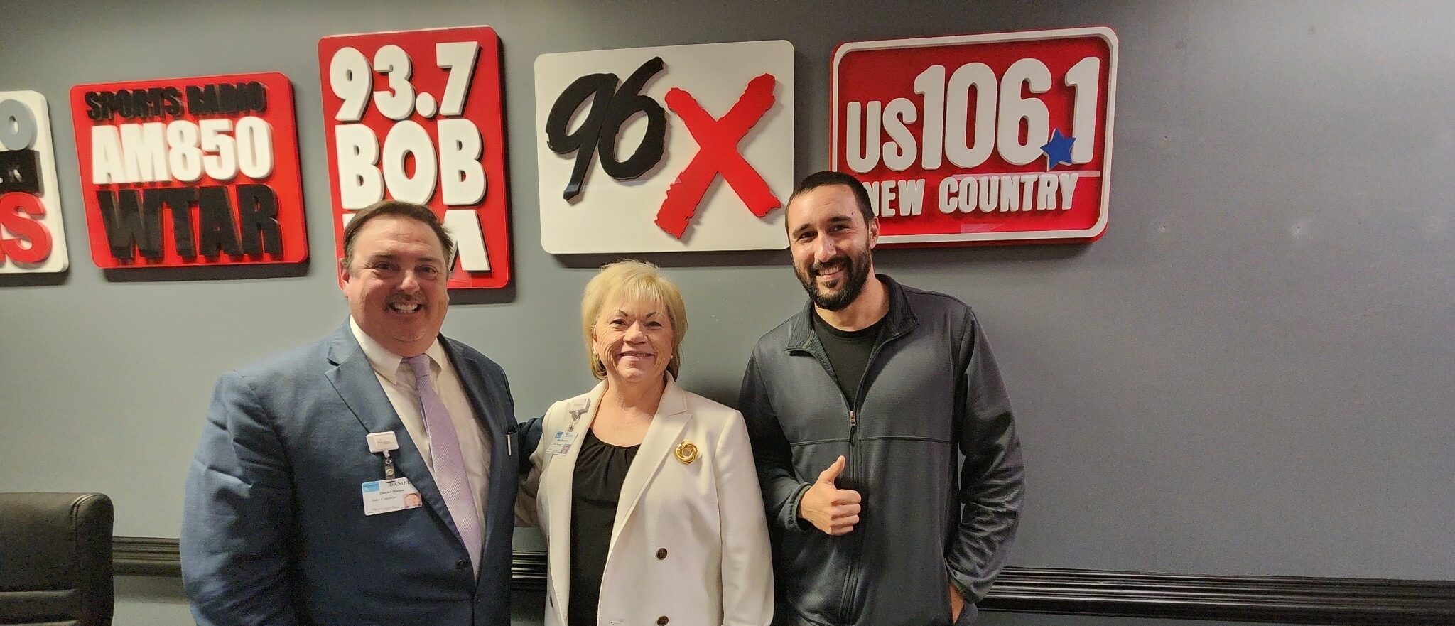 Three people standing in front of a wall with radio station signs. One person in a suit, another in a white jacket, and the third in casual attire giving a thumbs up.