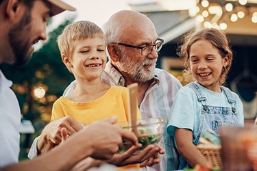 A joyful family gathering outdoors, featuring a bearded older man sitting with a young boy and girl. The boy wears a yellow shirt, and the girl wears denim overalls. They are smiling and sharing a meal. Bright lights are strung in the background.