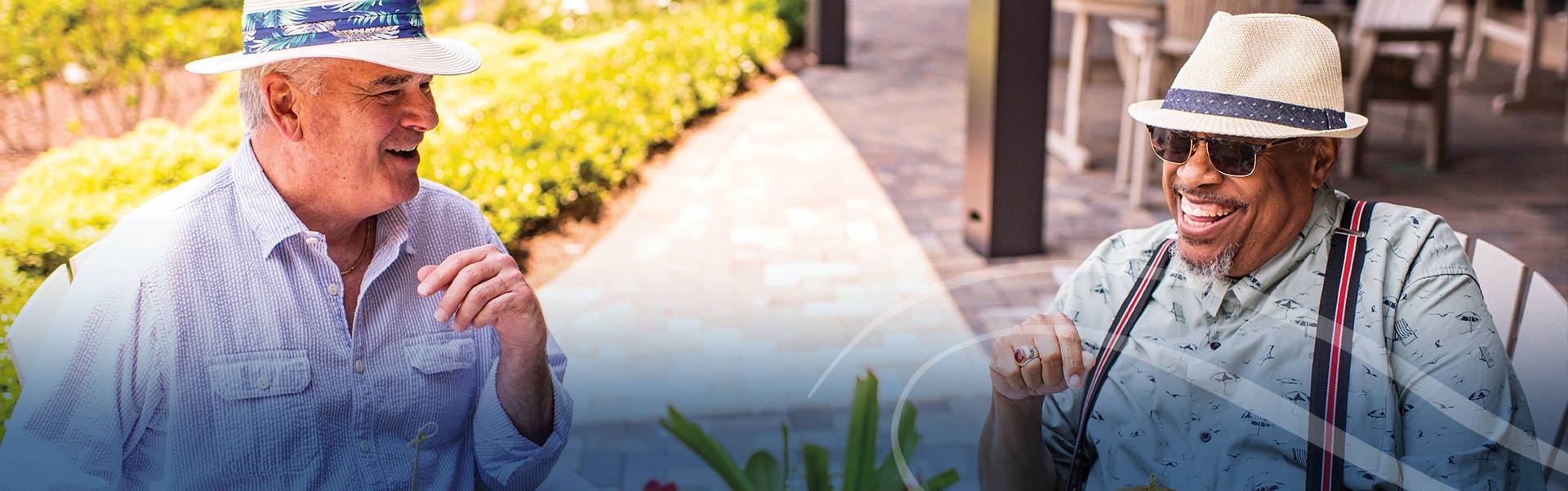 Two elderly men sit outdoors, laughing and enjoying a conversation. Both wear hats and sunglasses, one in a light shirt and the other in a patterned shirt with suspenders. The scene is sunny, with a blurred background of greenery and patio seating.