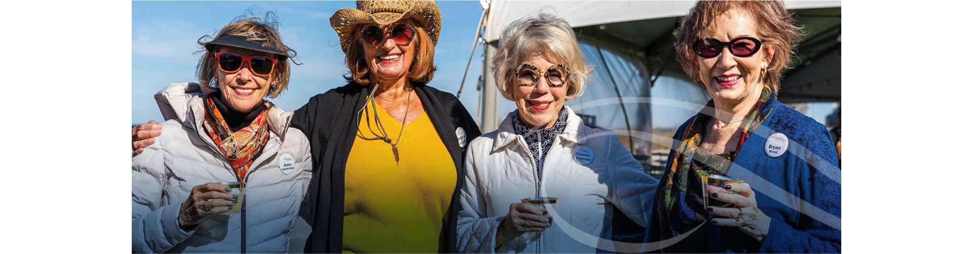 Four women, wearing sunglasses and stylish hats, smile as they hold drinks outdoors. They are dressed in bright, colorful clothing, suggesting a cheerful gathering under clear skies. A tent is visible in the background.