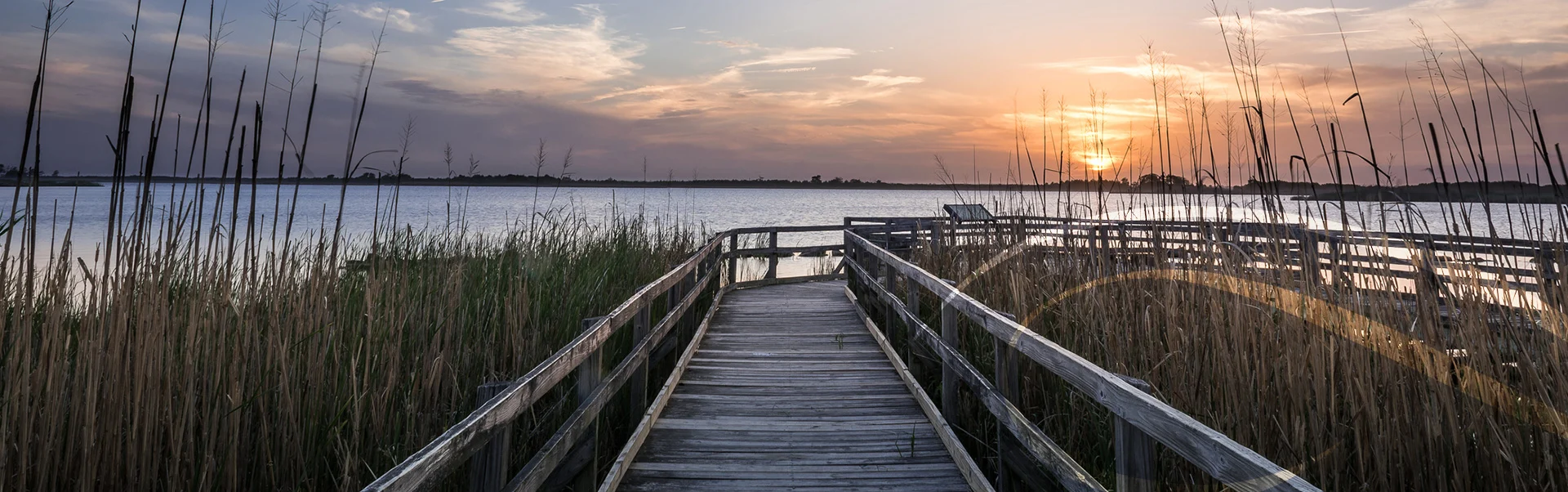 A wooden boardwalk extends through tall grasses toward a calm lake, with a vibrant sunset painting the sky in hues of orange and purple. The serene water reflects the colorful sky, creating a peaceful and picturesque scene.