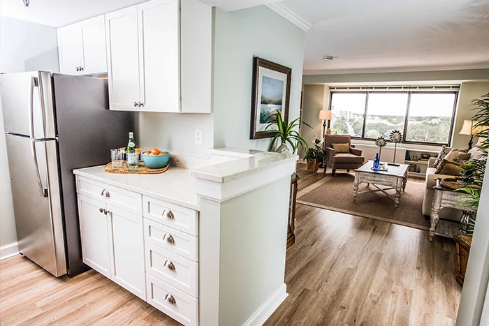 A modern open-plan living space with a white kitchen, light wood flooring, and a cozy living room. The kitchen has stainless steel appliances and a small counter with a plant. A large window offers a view of greenery in the background.