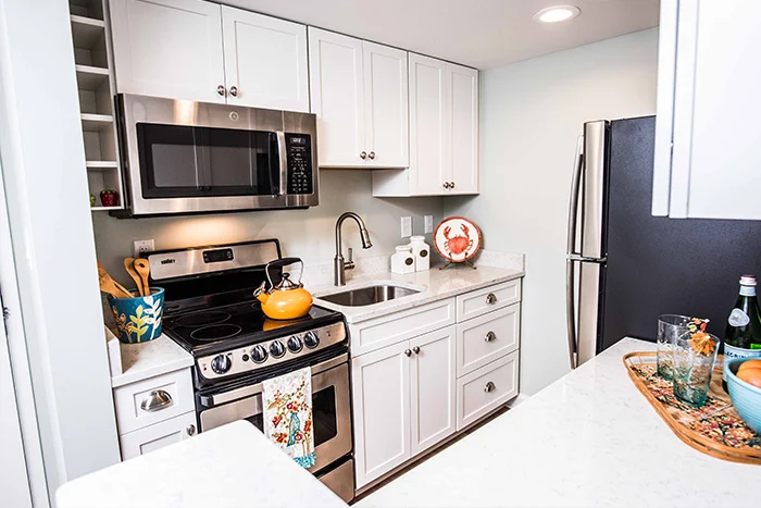 A modern kitchen with white cabinets, stainless steel appliances, and a marble countertop. Theres a stove, microwave, and a refrigerator. A colorful teapot sits on the stove, and a decorative plate is displayed on the counter.
