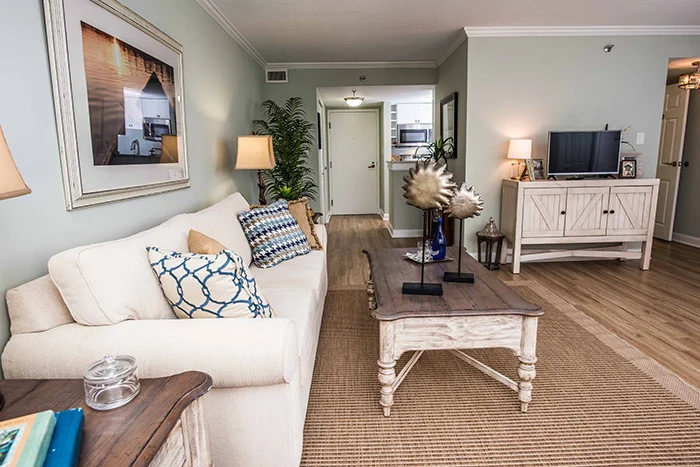 A cozy living room with light green walls features a white couch adorned with patterned cushions. A wooden coffee table sits on a beige rug, and a TV is on a cabinet nearby. A plant is in the corner, and artwork decorates the wall.