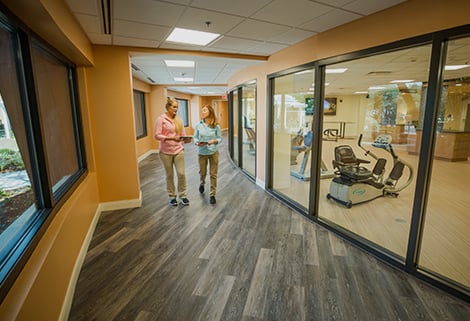 Two people walk down a curved hallway with large windows. They are engaged in conversation. The windows show a room with exercise equipment. The hallway has wooden floors and warm-toned walls, creating a welcoming environment.