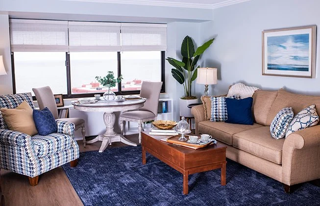 A cozy living room with a beige sofa, blue and white patterned armchair, and a wooden coffee table. A navy rug covers the floor. A round dining table with chairs sits by the window. A large potted plant and ocean-themed artwork complete the decor.