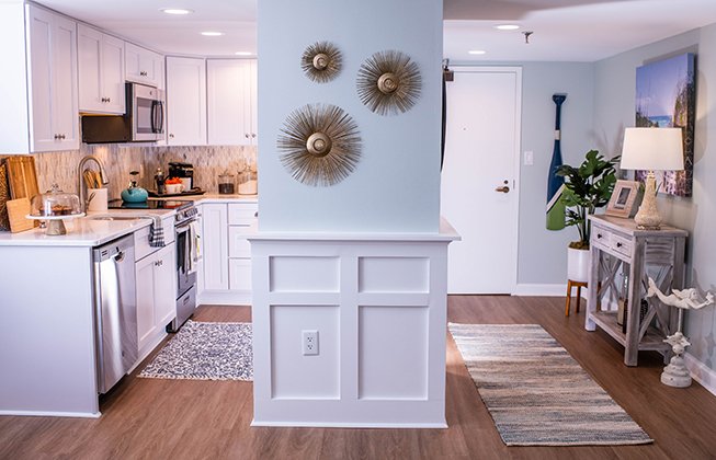 A modern kitchen and living area with white cabinets, stainless steel appliances, and wooden flooring. Decorative sunburst wall hangings and rugs add warmth. Plants and nautical decor are visible near the entryway.