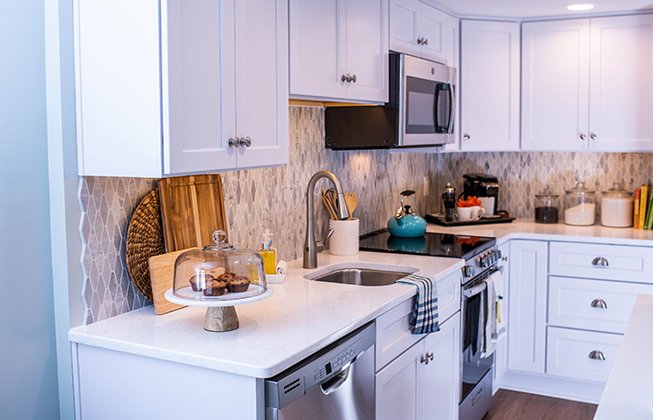 A modern kitchen with white cabinets, a stainless steel stove and microwave, and a patterned backsplash. A cake stand with cookies is on the counter next to a sink. Various kitchen items are neatly arranged on the countertop.