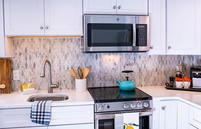Modern kitchen with white cabinets, gray patterned backsplash, and stainless steel appliances. A turquoise kettle sits on the stove. A sink with a silver faucet is on the left, and cooking utensils are in a holder on the countertop.