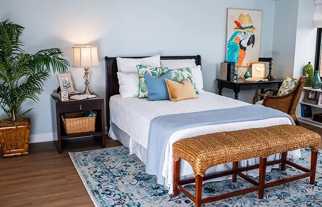 A cozy bedroom with a bed covered in white and blue linens, decorative pillows, and a woven bench at the foot. A bedside table with a lamp and a potted plant are nearby. Art decorates the light blue walls, enhancing the tranquil atmosphere.