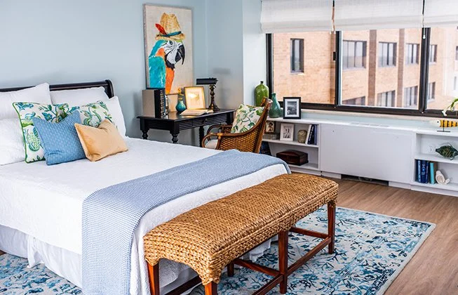 A cozy bedroom with a bed featuring white and blue bedding, a woven bench at the foot, and a parrot painting on the wall. A window overlooks a cityscape, and theres a wicker chair and a small desk with framed photos and books.