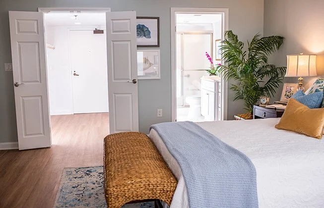 A cozy bedroom features a neatly made bed with white and blue bedding. A woven bench sits at the foot of the bed. A potted plant and a lamp are on the bedside table. Open doors lead to a hallway and bathroom with a plant decoration.