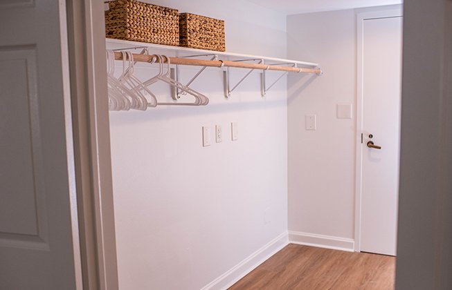 A minimalistic closet with wooden flooring, a white wall, and an open white door. It features a wooden shelf with woven baskets and empty hangers on a metal rod.