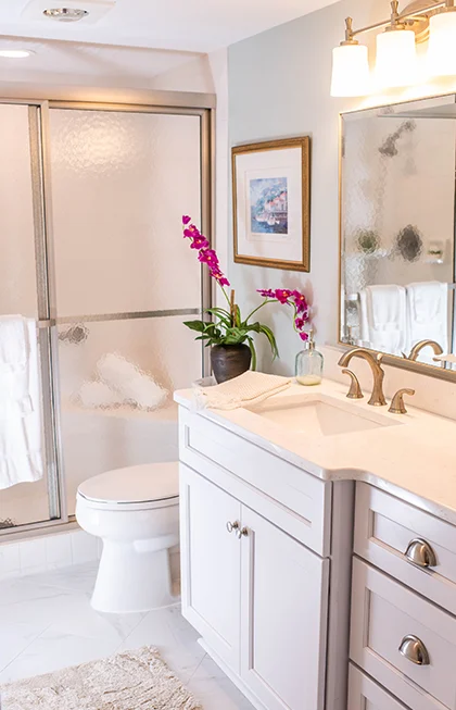 A bright bathroom featuring a white vanity with a gold faucet, a mirror, and a potted orchid on the counter. A glass-enclosed shower with towels is in the background, and artwork hangs on the wall. The room is well-lit with modern light fixtures.