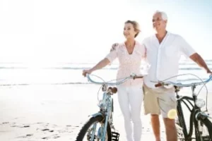 An older couple walks their bicycles on a sunny beach, smiling and enjoying the day. The ocean is visible in the background under a clear sky.