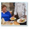 A chef serves a seated woman a dish of pasta with vegetables. The woman, wearing a blue sweater, is smiling. A menu and silverware are on the table.