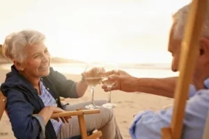An elderly couple sitting in wooden chairs on a beach clink wine glasses, smiling at each other. The sun is setting in the background, casting a warm glow over the scene.