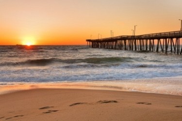 Sunset over a beach with gentle waves and a sandy shore. A pier extends into the ocean from the right, and the sun casts a warm, orange glow across the sky and water. Footprints are visible in the sand in the foreground.
