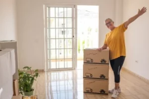 A person in a yellow shirt and black pants stands happily next to stacked cardboard boxes in an empty room. The room has tiled floors, large windows, and a potted plant on the floor. Sunlight streams in through the windows.