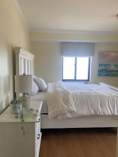 A bright bedroom with a white bed, fluffy comforter, and a white headboard. A small white nightstand holds a lamp and plant. A window with blinds lets in natural light, and a colorful painting is visible on the wall. The floor is wooden.