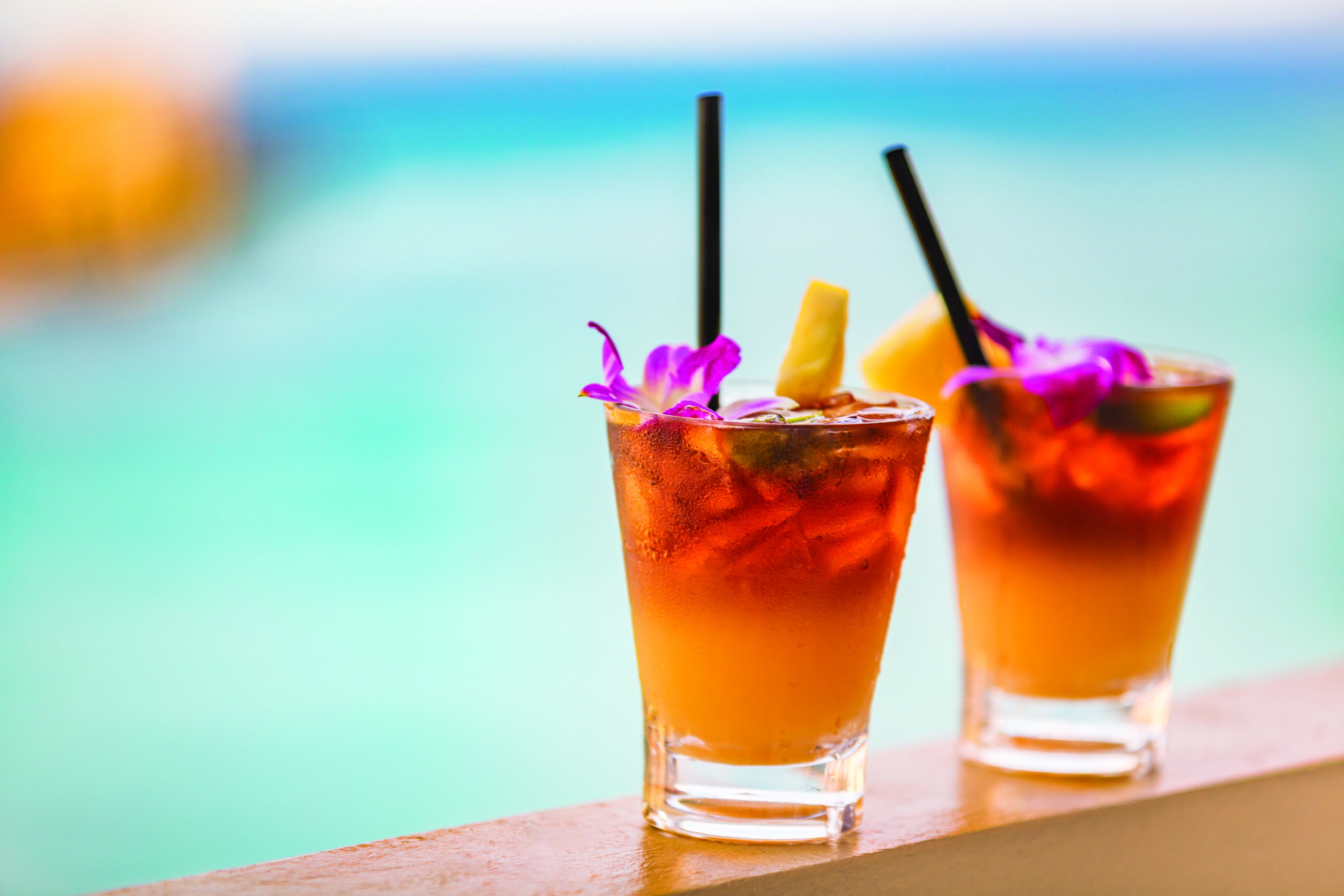 Two tropical cocktails with ice, garnished with purple flowers, pineapple slices, and straws, sit on a ledge overlooking a turquoise ocean backdrop.