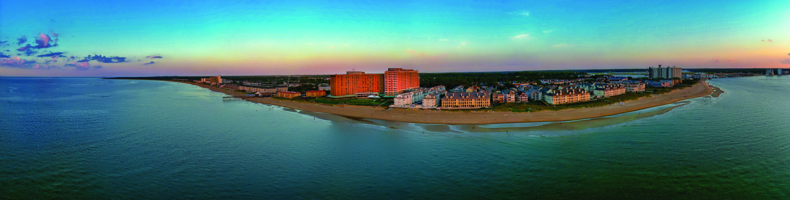 A panoramic view of a coastal town at sunset, with buildings lining a sandy beach bordered by calm ocean waters. The sky is a gradient of blue to orange, creating a serene and picturesque scene.