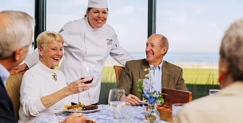 A joyful group of people sit around a dining table, smiling and engaged in conversation with a chef standing beside them. The table is set with plates of food and glasses of wine, and a scenic ocean view is visible through the large windows behind them.