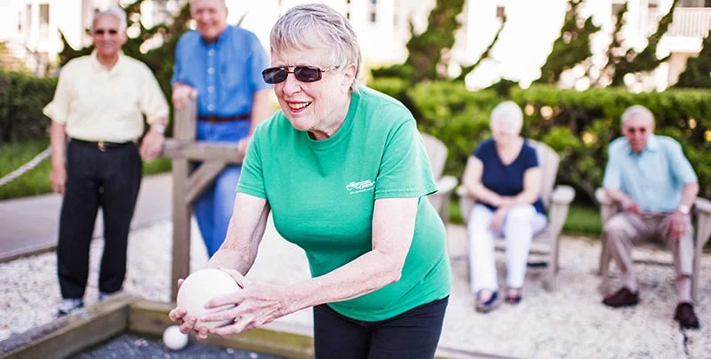 An older woman in a green shirt and sunglasses prepares to throw a bocce ball. Four other people sit and stand in the background, watching her play. The setting is outdoors on a sunny day.