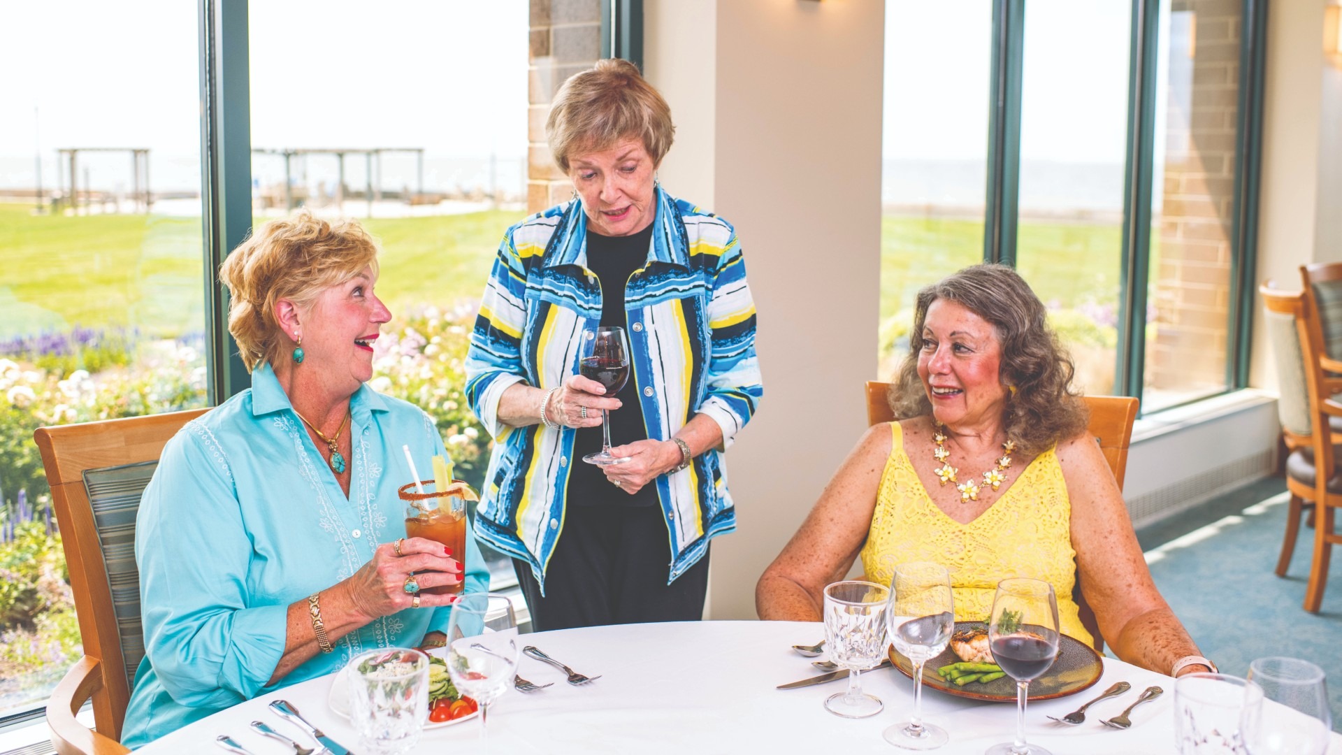 Three women are enjoying a meal together at a table by large windows with a scenic view. One woman stands holding a glass, while the other two are seated, engaging in conversation and smiling. The table is set with drinks and plates of food.